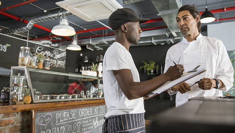 Restaurant worker talking with chef, taking notes on clipboard